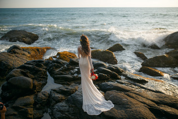 Beautiful bride stands on a cliff above the sea in a glamorous white wedding dress with a train