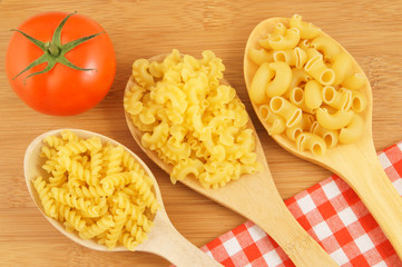 Macaroni on wooden and tablecloth background with spoons and tomato