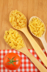 Macaroni on wooden and tablecloth background with spoons and tomato