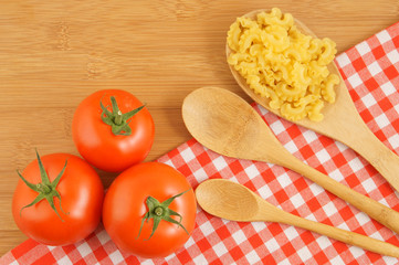 Macaroni on wooden and tablecloth background with spoons and tomatoes