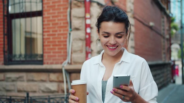 Slow motion of beautiful lady using smartphone holding to go coffee walking outdoors smiling in the street. People, devices and moden lifestyle concept.