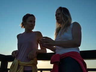 Two young girls in sportswear are standing on the promenade, leaning on the railing and talking. The rising sun shines brightly.