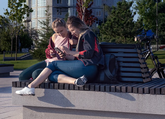Two young girls in sportswear are sitting on a bench in a city square and listening to music. They are holding phones in their hands. Girls rest after a bike ride.