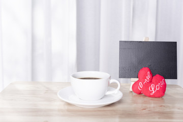 Hot coffee cup with red hearts shape symbol  and black board on wood table  with white curtain background