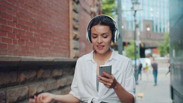 Slow motion of pretty woman listening to music in headphone singing using smartphone outdoors walking alone having fun. People, devices and city lifestyle concept.