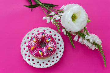 Donut with bright sugar pink icing on a white openwork plate on a pink purple table, where there are white flowers and a place for text.