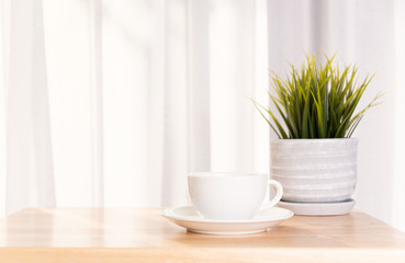 White coffee cup and plant pot on wooden office desk  with morning sunlight and  soft curtain background