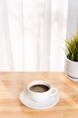 White coffee cup and plant pot on wooden office desk  with morning sunlight and  soft curtain background