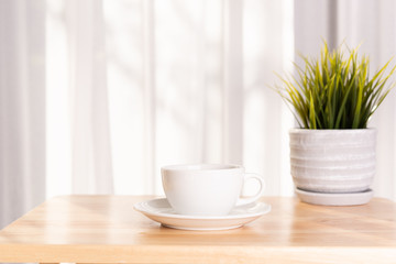White coffee cup and plant pot on wooden office desk  with morning sunlight and  soft curtain background