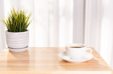White coffee cup and plant pot on wooden office desk  with morning sunlight and  soft curtain background