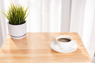 White coffee cup and plant pot on wooden office desk  with morning sunlight and  soft curtain background