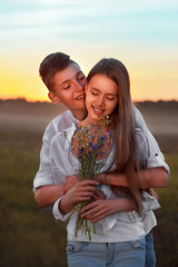 A young guy gives wild flowers to a beautiful girl in the evening at sunset against a background of rural nature. Romantic relationship between two young people. First love or friendship.