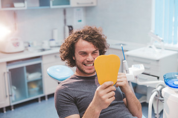 Charismatic smiling young man on the dentist chair , after a oral hygiene procedure take a mirror and looking at his white healthy teeth and are very happy for results , modern dental clinic