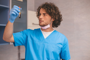 Fototapeta na wymiar Young doctor in front of the camera wearing a mask and surgical blue gloves holding a blood test tub and looking straight to the camera with a large smile