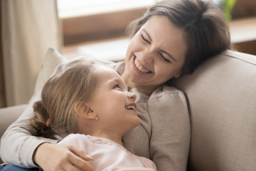Preschool daughter and cheerful mother lying on couch joking laughing