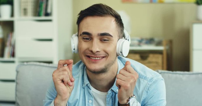 Caucasian attractive young hapy guy sitting on the sofa and resting while enjoying music via headphones at home. Portrait.