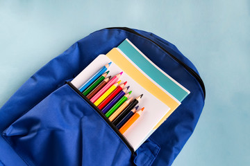 Multicolored wooden pencils and notebooks in a backpack on a paper blue background with copy space. School accessories. Top view