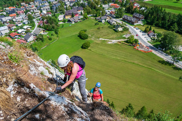 Women on a via ferrata route on Grancisce (Grancise) hill, above the Mojstrana village in Slovenia, on a sunny day, with tiny houses visible below. Outdoor adventures concept in Slovenia.