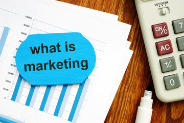 Text sign showing what is marketing. The text is written on a small wooden board. Graphs on the paper sheet, markers, keyboard, wooden background are on the photo.