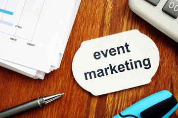Text sign showing event marketing. The text is written on a small wooden board. Graphs on the paper sheet, pen, keyboard, wooden background are on the photo.