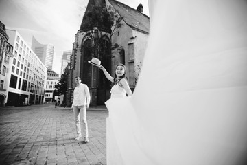 Happy young couple walking near old church in Leipzig. Pregnant woman in beautiful white dress throws up her hat