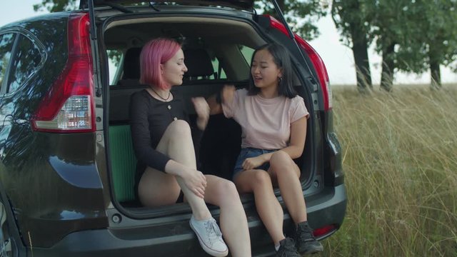 Joyful multiracial female friends playing rock paper scissors game while sitting in car trunk during summer vacations. Cheerful young women having fun playing hand game in car during road trip.