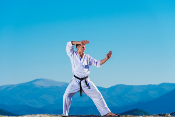 Blond karate athlete does kata on top of a mountain while performing a line up of kicks, punches and blocks on top of a mountain on a sunny day.