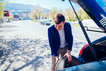 A tired businessman on a sunny day is taking a tire out of his car in order to be able to change his flat tire.