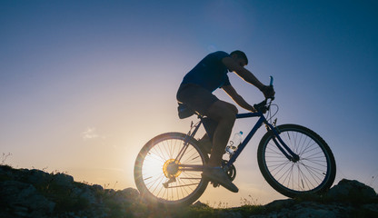 Fototapeta na wymiar Silhouette of a fit male mountain biker riding his bike uphill on rocky harsh terrain on a sunset.