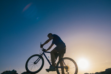 Naklejka na ściany i meble Silhouette of a fit male mountain biker riding his bike uphill on rocky harsh terrain on a sunset.