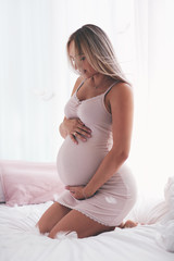 beautiful tender pregnant woman sitting on the bed in the morning, holding hands on belly, maternity concept