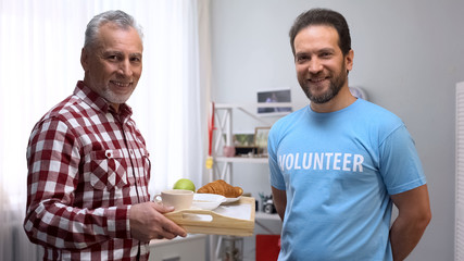 Middle aged volunteer and senior man looking at camera, holding tray with food