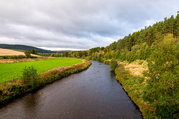 Fototapeta na wymiar A view of the flowing river with a forest on one side and agricultural lands on the other side, autumn in Cairngorms National Park