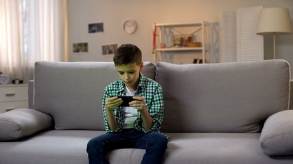 Obraz premium Preschool boy procrastinating and playing game on smartphone, gadget addiction