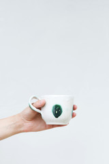 White ceramic mug in a female hand on a background of a white wall. Hand-made