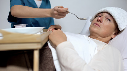 Terminally ill woman looking at camera, nurse feeding patient, Alzheimer disease