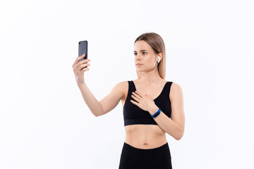 Young sporty blond woman in a black sportswear making selfie with smartphone showing bicep after workout standing over white background. Model listens music during workout with wireless headphones