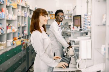 Fototapeta premium Two young cheerful pharmacists African man and Caucasian woman working together and using computers. Pharmacists searching medicines at pharmacy