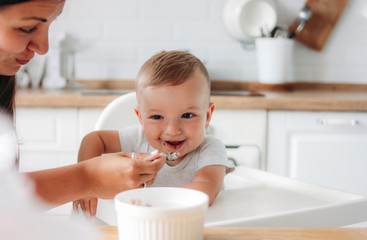 Charming little baby boy eating first food porridge from spoon at kitchen. Mom feeds child