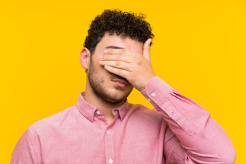Man with curly hair over isolated yellow wall covering eyes by hands