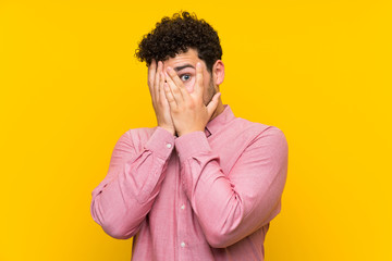 Fototapeta na wymiar Man with curly hair over isolated yellow wall covering eyes and looking through fingers