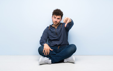 Young man sitting on the floor showing thumb down with negative expression