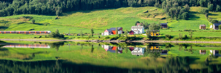 Fototapeta na wymiar Train passing a traditional Norwegian summer farm along lake Vangsvatnet reflected in the water near Voss Hordaland county Norway
