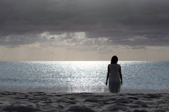 Woman silhouette from behind dressed in a light white dress walks sadly and thoughtfully towards the dark sea at dawn with dramatic sky