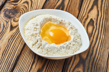 Broken egg in ceramic bowl with flour lies on rustic wooden table