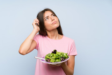 Young woman with salad over isolated blue background having doubts and with confuse face expression