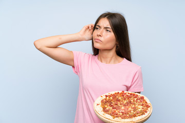Pretty young girl holding a pizza over isolated blue wall having doubts and with confuse face...