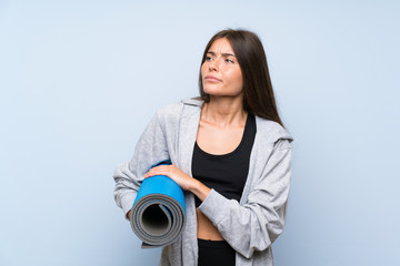 Young sport girl with mat over isolated blue background making doubts gesture while lifting the...