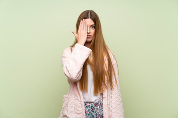 Young woman in dressing gown over green wall covering a eye by hand