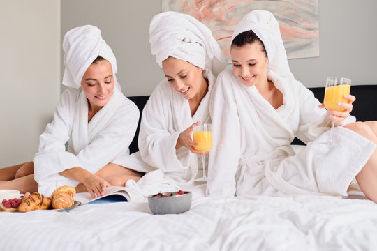 Young beautiful women in white bathrobes and towels on head dreamily reading book together. Pretty girls holding glasses with orange juice in hands having breakfast in bed in hotel - Powered by Adobe